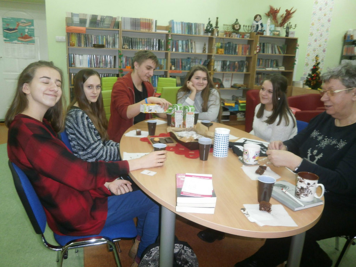 Wnętrze biblioteki. Przy stoliku siedzą uczestnicy DKK SP2 dyskutują o książce  "Błysk flesza" o problemach młodzieży.Na stoliku soki i ciastka.