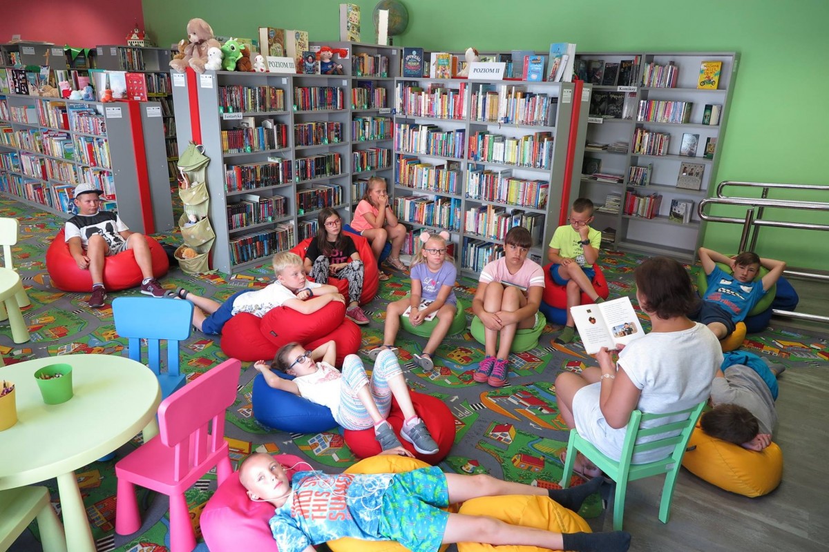 Wnętrze biblioteki. Na pufach siedzą dzieci słuchają opowiadania o przygodach Mikołajka i jego zabawnych przyjaciół, czyta bibliotekarka Ania Wiśniewska.