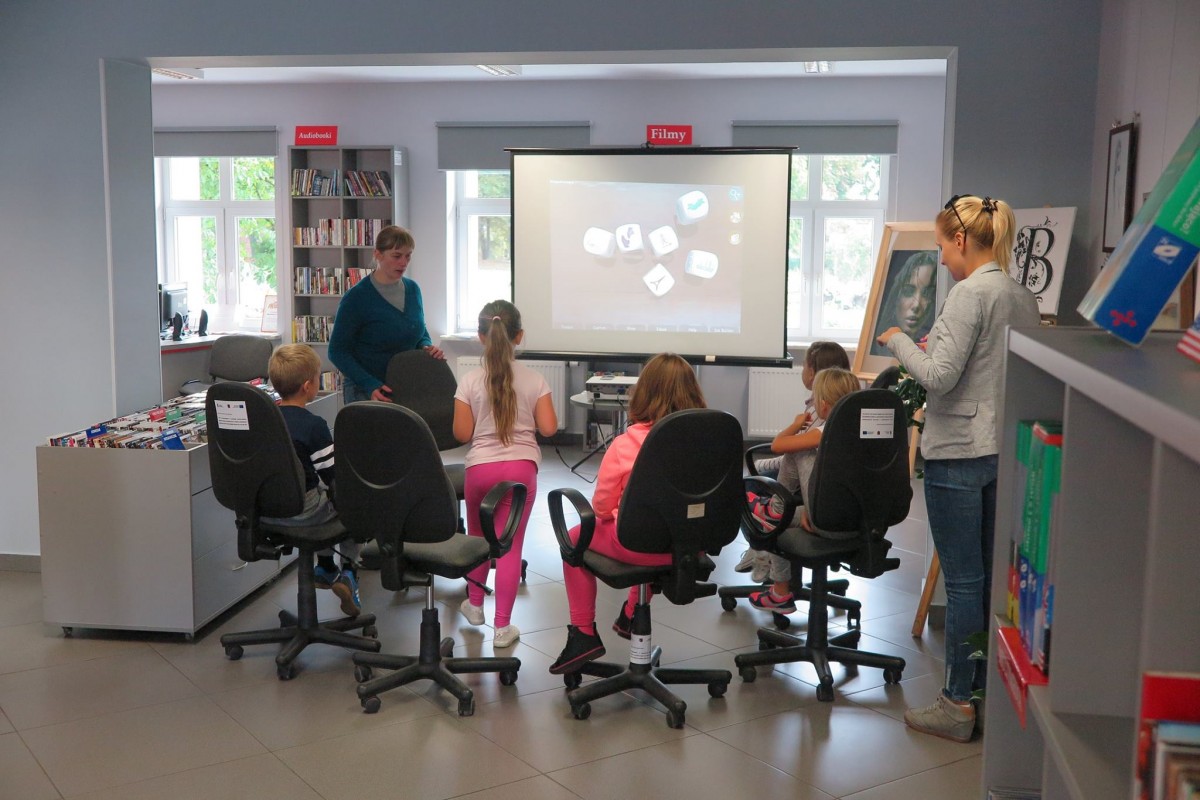 Wnętrze biblioteki,mediateka. Na krzesełkach siedzą dzieci biorą udział w warsztatach z programowania z wykorzystaniem ozobotów oraz kostek do storytellingu. W tle monitor z prezentacją. 