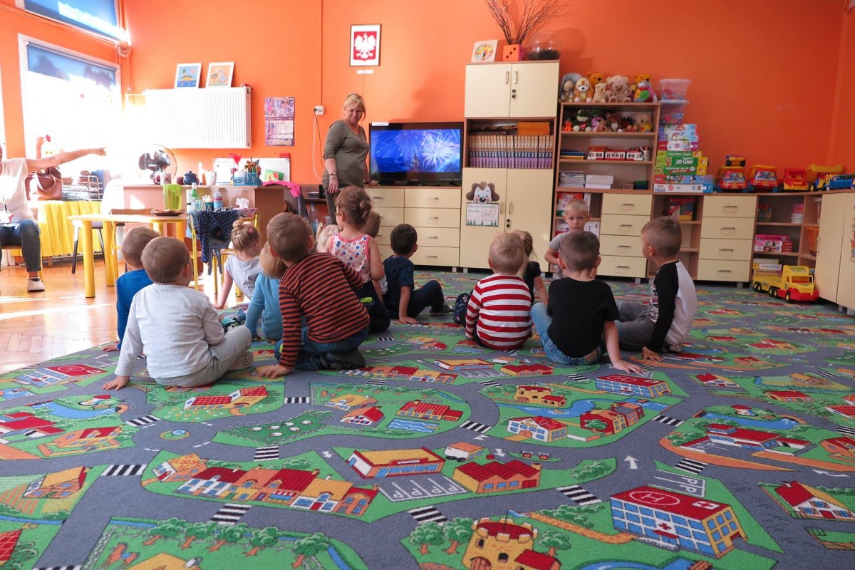 Wnętrze Przedszkola nr 5 w Działdowie. Na kolorowej wykładzinie siedzą dzieci i oglądają bajkę, w tle telewizor. 