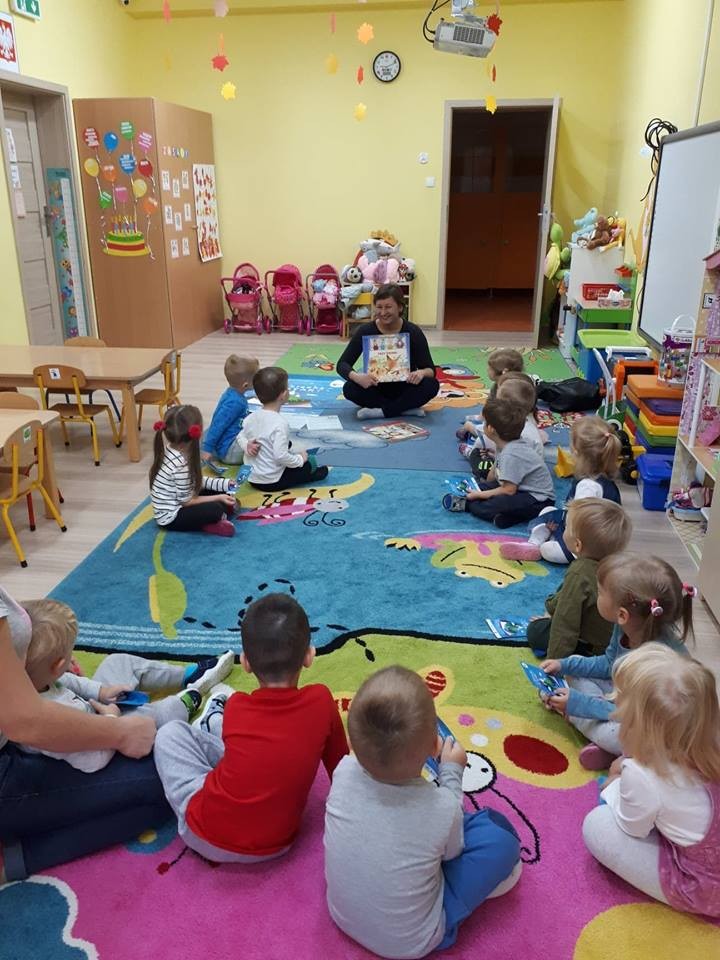 Wnętrze Przedszkola ''U Misia'' , na kolorowej wykładzinie siedzą dzieci , zajęcia prowadzi bibliotekarka Ania Wiśniewska.