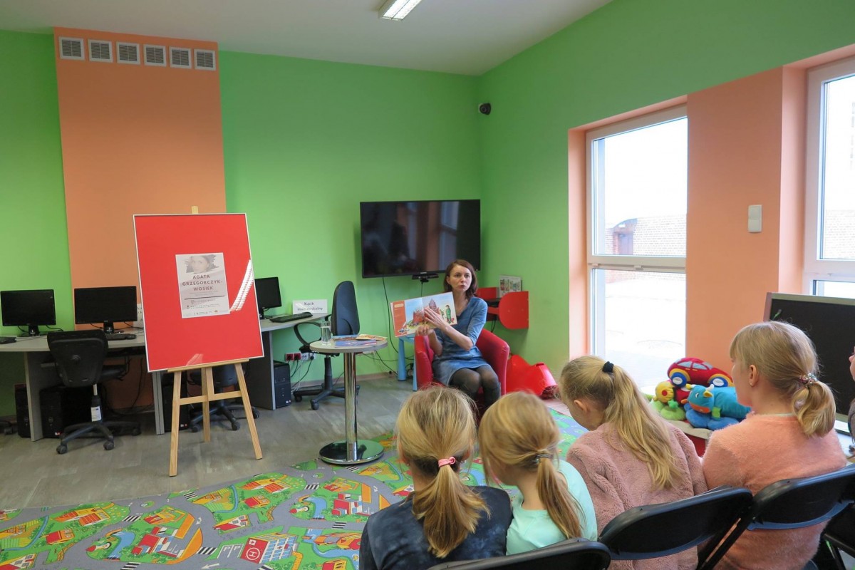 Wnętrze biblioteki. Na czerwonym fotelu siedzi autorka bajek dla dzieci, olsztynianka z urodzenia, Pani Agata Grzegorczyk – Wosiek pokazuje swoją książkę dzieciom.
