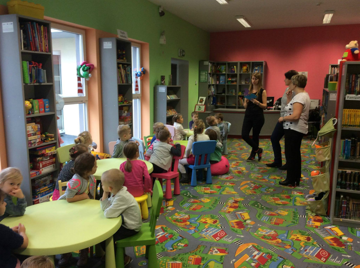 Wnętrze biblioteki, przy stolikach siedzą dzieci, bibliotekarka Aleksandra Cybulska prowadzi zajęcia edukacyjne. 