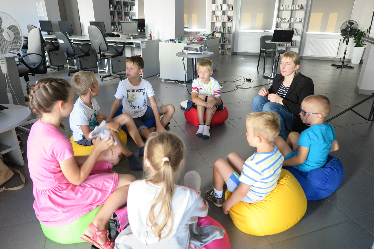 Wnętrze biblioteki,mediateka. Na pufach siedzą dzieci z bibliotekarką Małgorzatą Trąmpczyńską wszyscy biorą udział w zajęciach z cyklu „nowe technologie”.