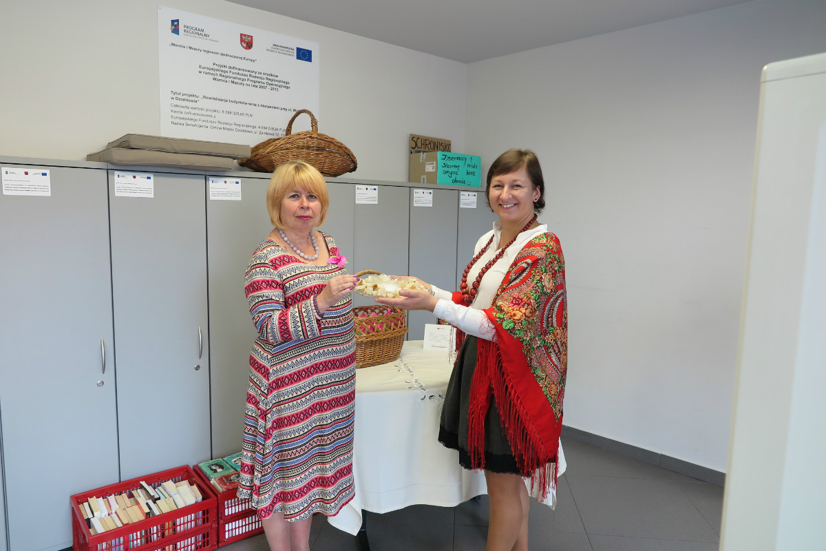 Wnętrze biblioteki. Bibliotekarka Ania Wiśniewska z okazji Narodowego Czytania częstuje chlebem i solą panią Elżbietę Zakrzewską.
