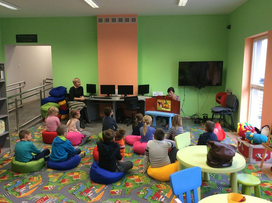 Wnętrze biblioteki. Grupa dzieci ze SP nr 3 słucha teatrzyku Kamishibai "Trzy świnki".