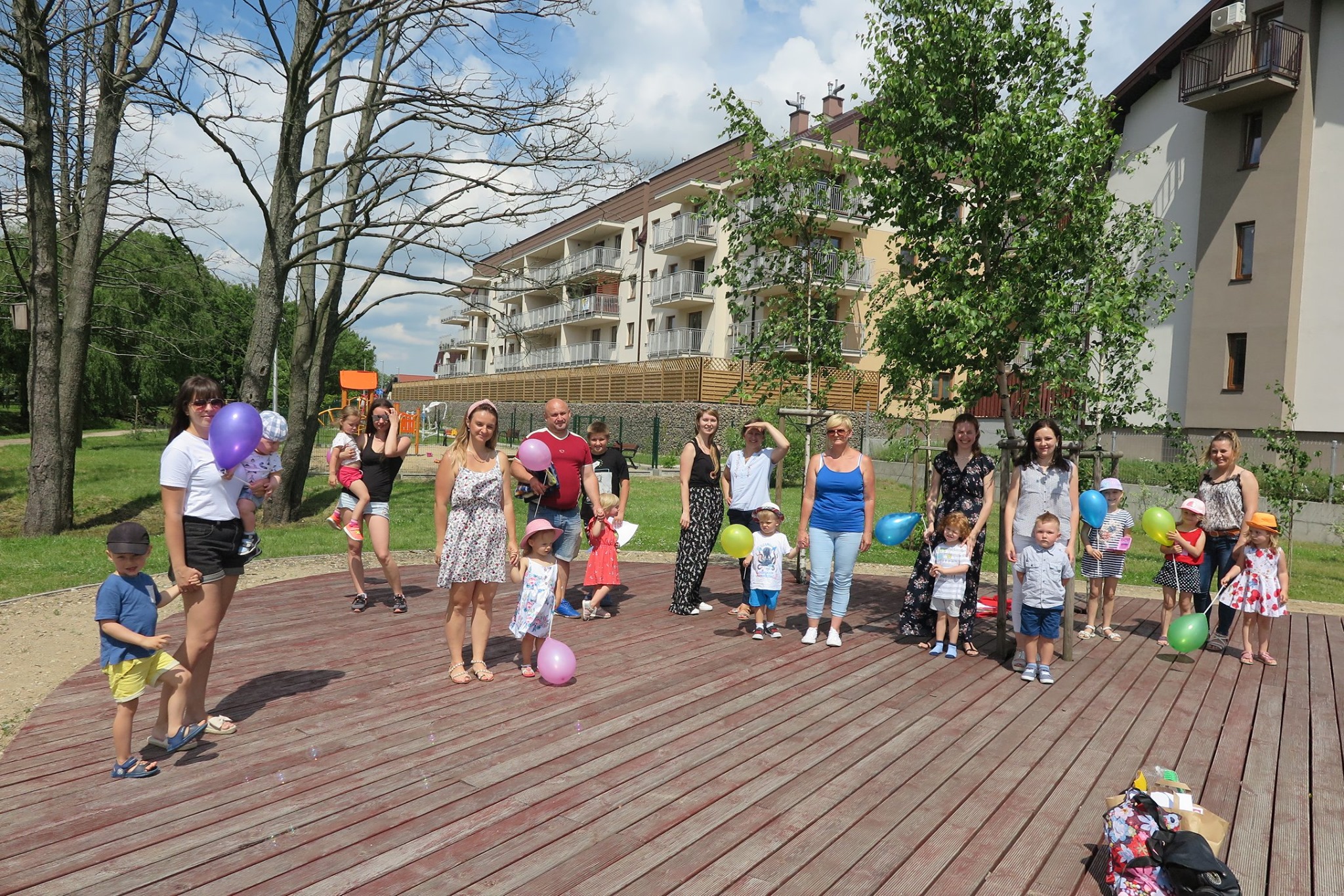 W parku bibliotekarka Aleksandra Cybulska zorganizowała spotkanie Klubu Malucha. 12 dzieci wraz z opiekunami uczestniczą w spotkaniu. Dzieci trzymają w ręku balony.