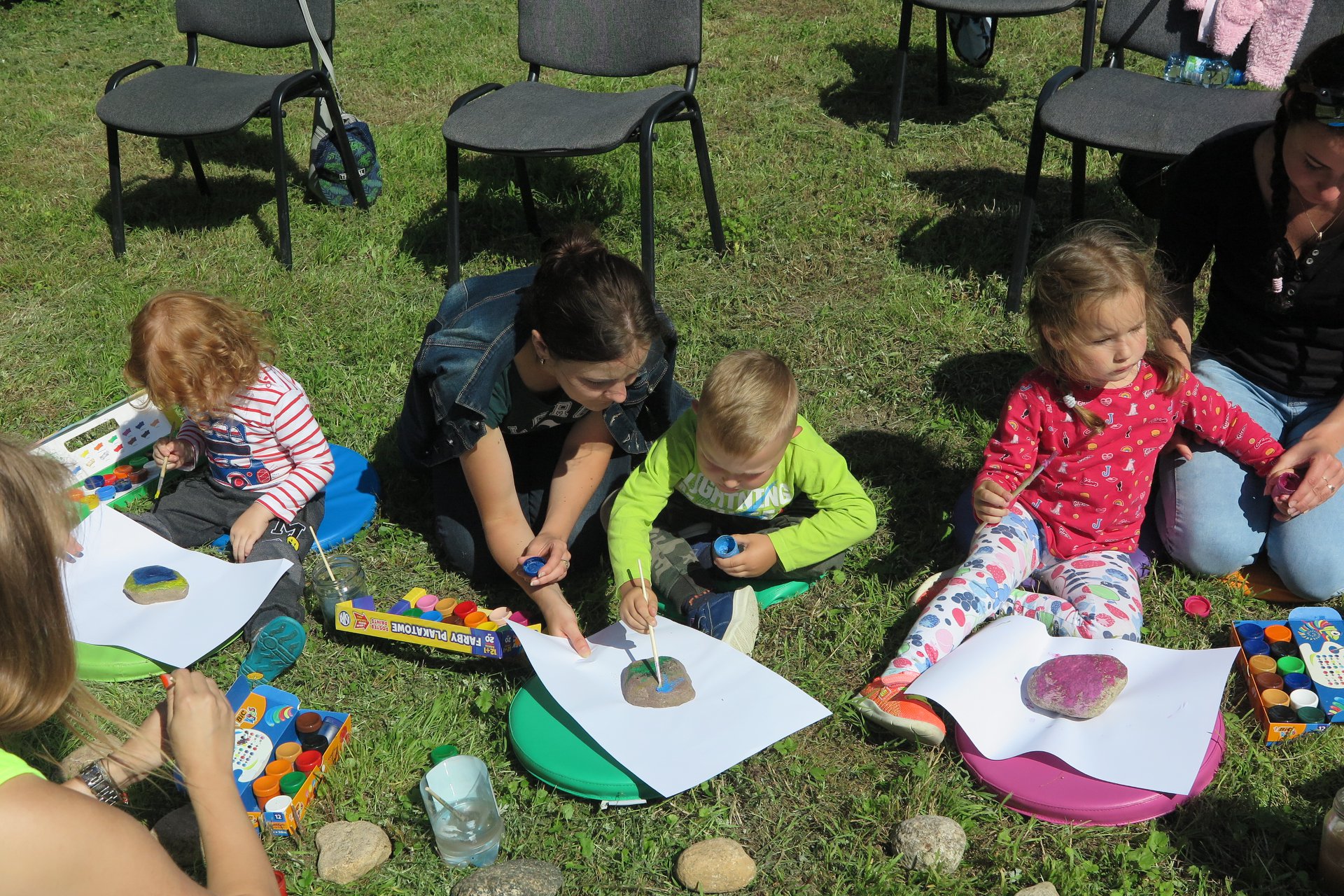 Na trawniku siedzi troje dzieci oraz dwóch opiekunów. Dzieci malują kamienie farbkami. 