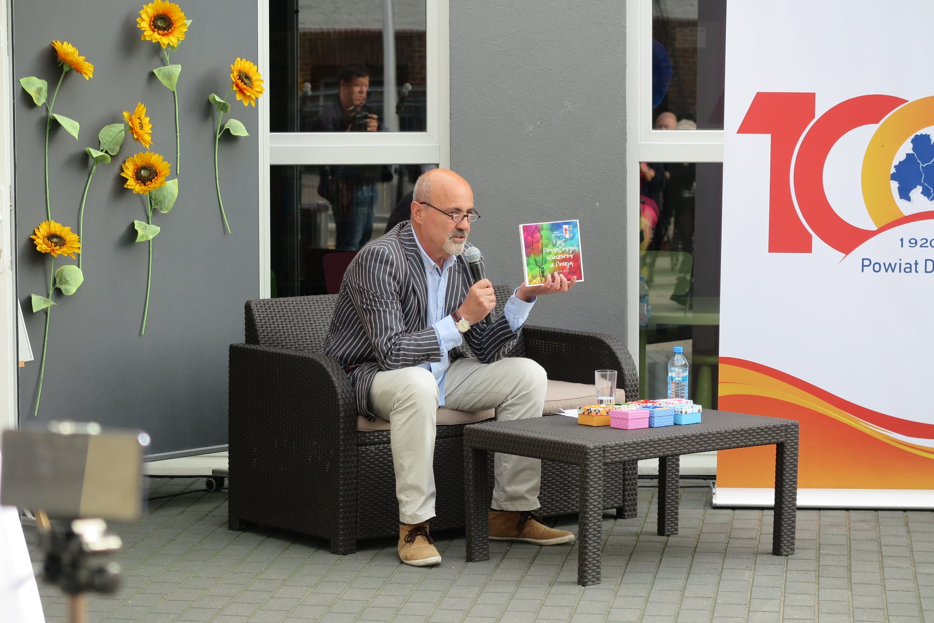 Na brązowej kanapie przed biblioteką siedzi aktor Marcin Troński w ręku trzyma mikrofon a w drugiej dłoni prezentuje książkę. 