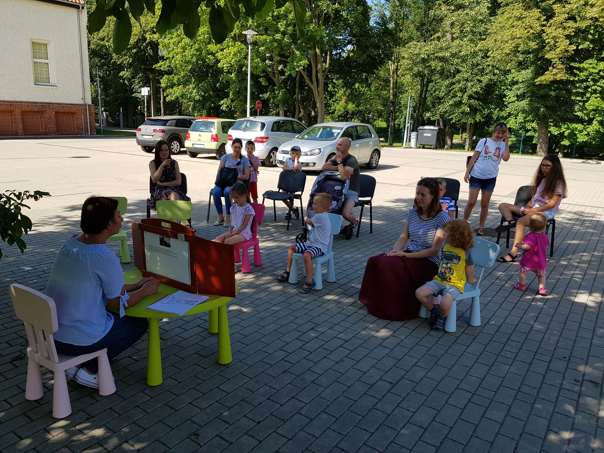 Bibliotekarka Ania Wiśniewska przy stoliku czyta dzieciom teatrzyk ''Słowik''. Na krzesełkach siedzą małe dzieci wraz z opiekunami. 