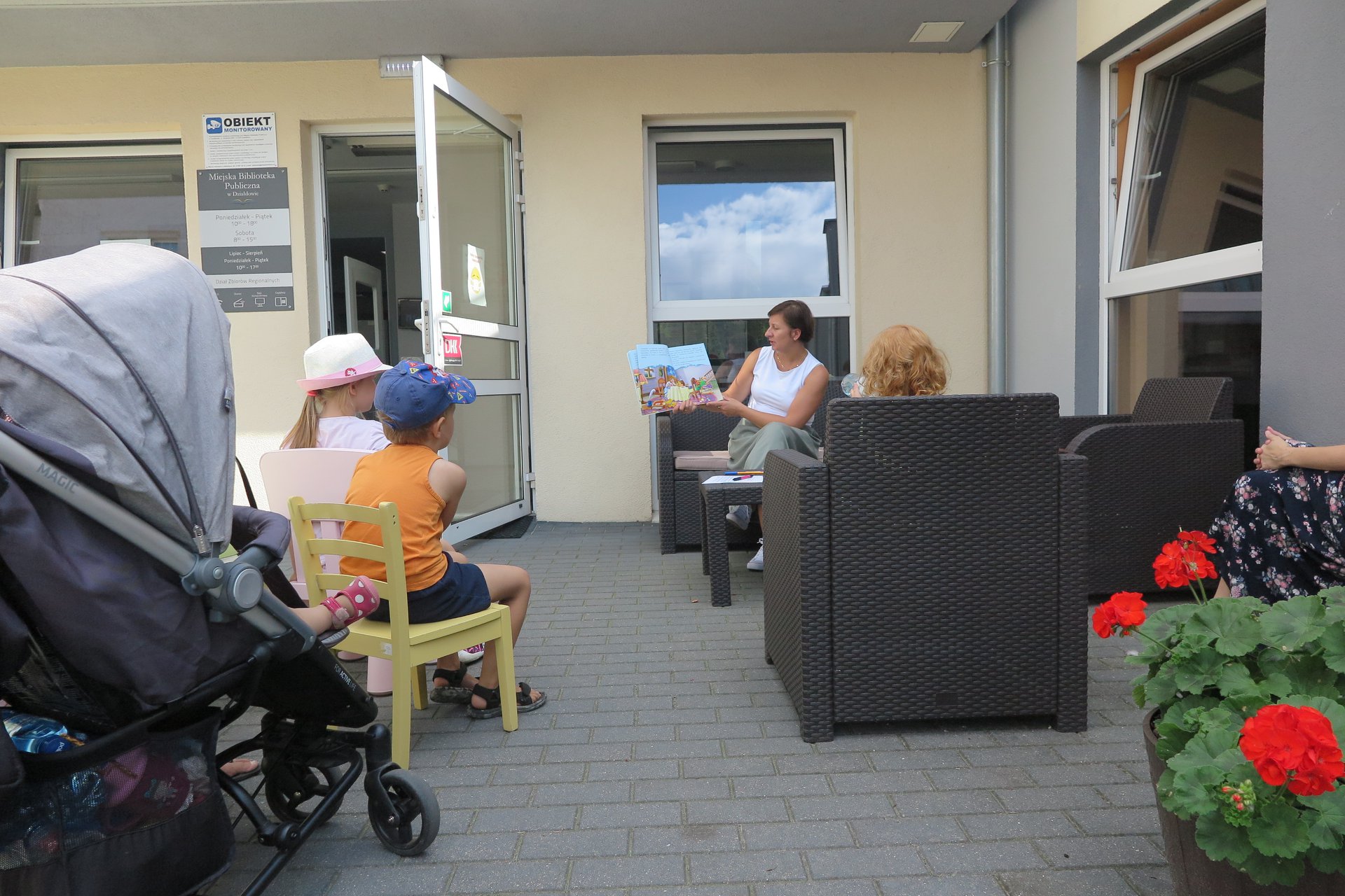 Spotkanie z książką podczas wakacji przed budynkiem biblioteki w Działdowie. Bibliotekarka Ania Wiśniewska czyta opowiadanie ,,Złotowłosa i trzy niedźwiadki''. Słuchają zgromadzone małe dzieci wraz z opiekunami. 
