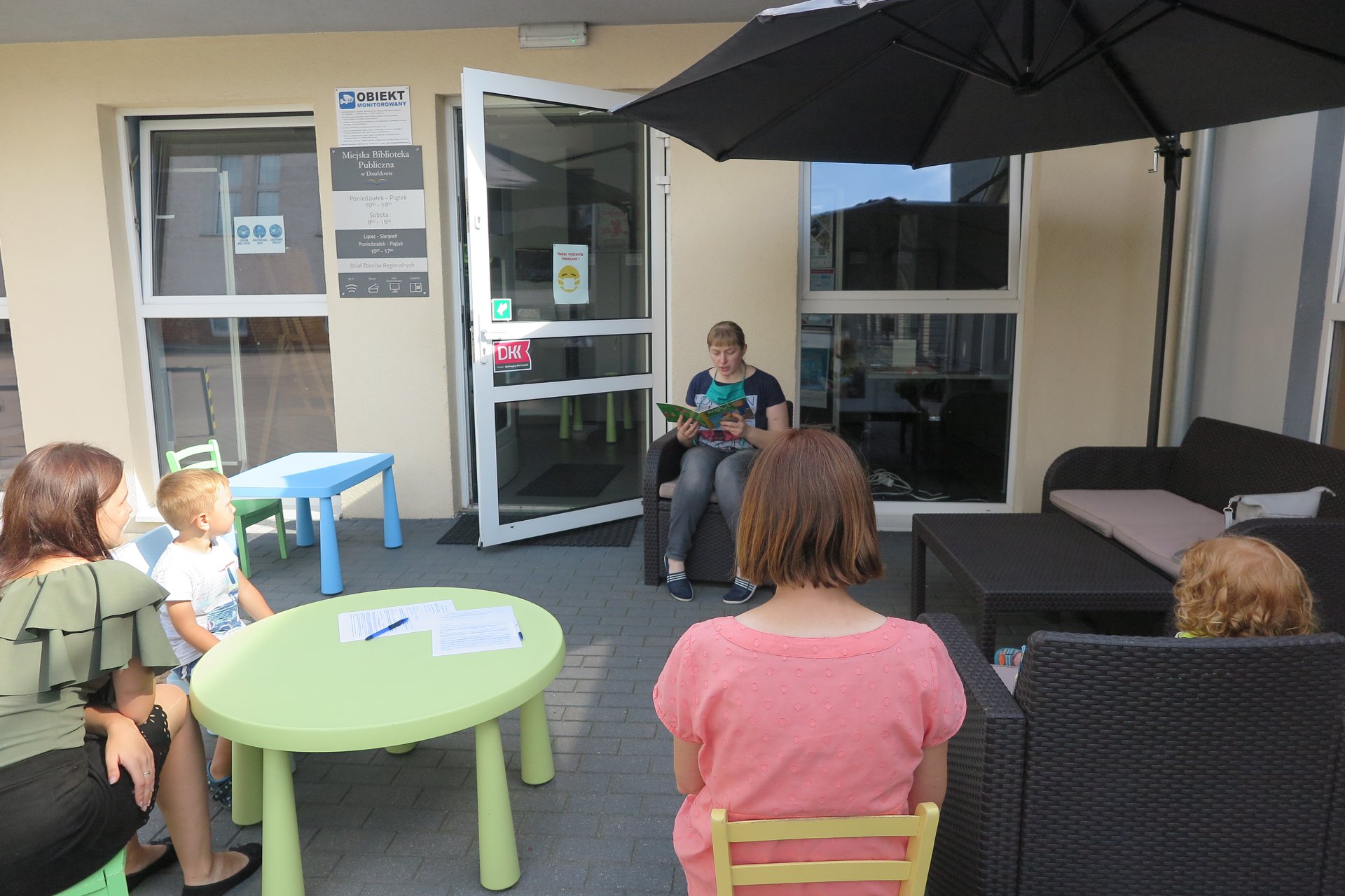 Letnie czytanie przed MBP w Działdowie. Bibliotekarka Małgorzata Trąmpczyńska pod parasolem czyta małym dzieciom. Dwoje dzieci siedzi na krzesłach wraz z nimi dwóch opiekunów. 
