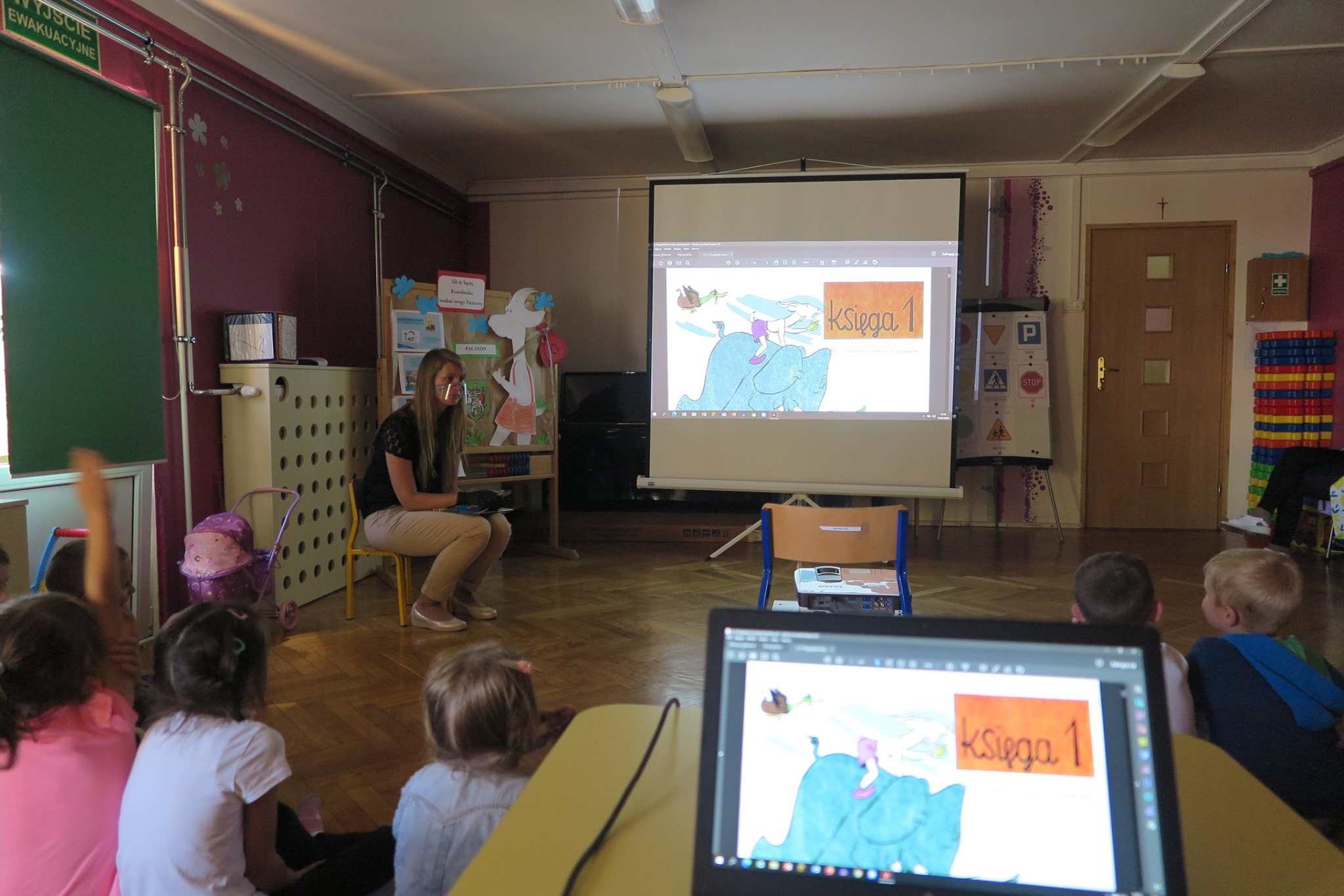 W tle monitor z bajką Koziołek Matołek obok bibliotekarka Aleksandra Cybulska prowadzi projekt edukacyjny z dziećmi z grupy IX ''Misie'' z Przedszkola nr 4 oraz dzieci.