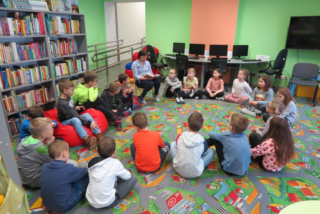 Wnętrze biblioteki. W kole na pufach siedzą dzieci i słuchają opowiadania,czyta bibliotekarka Ania Wiśniewska.