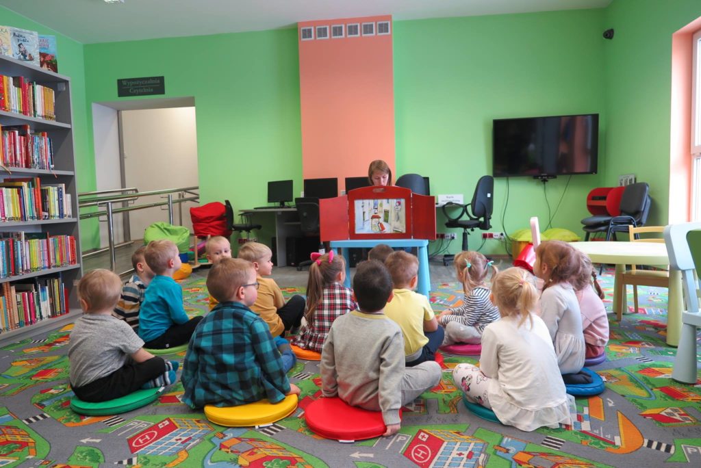 Wnętrze biblioteki.  Na pufach siedzą dzieci i słuchają teatrzyku Kamishibai "Pani Zamieć" , czyta bibliotekarka Ola Cybulska.