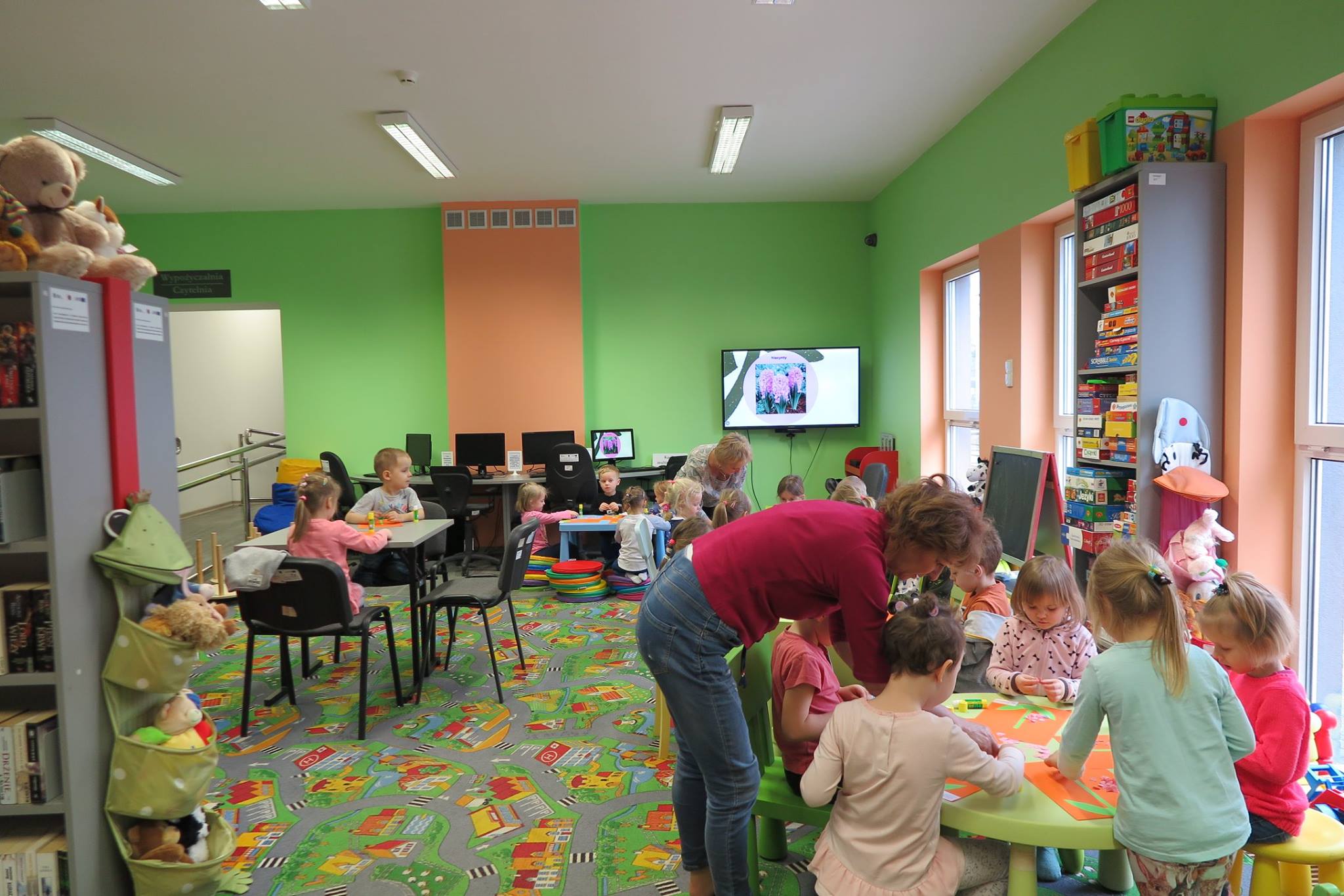 Wnętrze biblioteki. Przy stolikach siedzą dzieci i wykonują piękne kolorowe hiacynty z przygotowanych elementów, które potem zabiorą ze sobą do przedszkola.