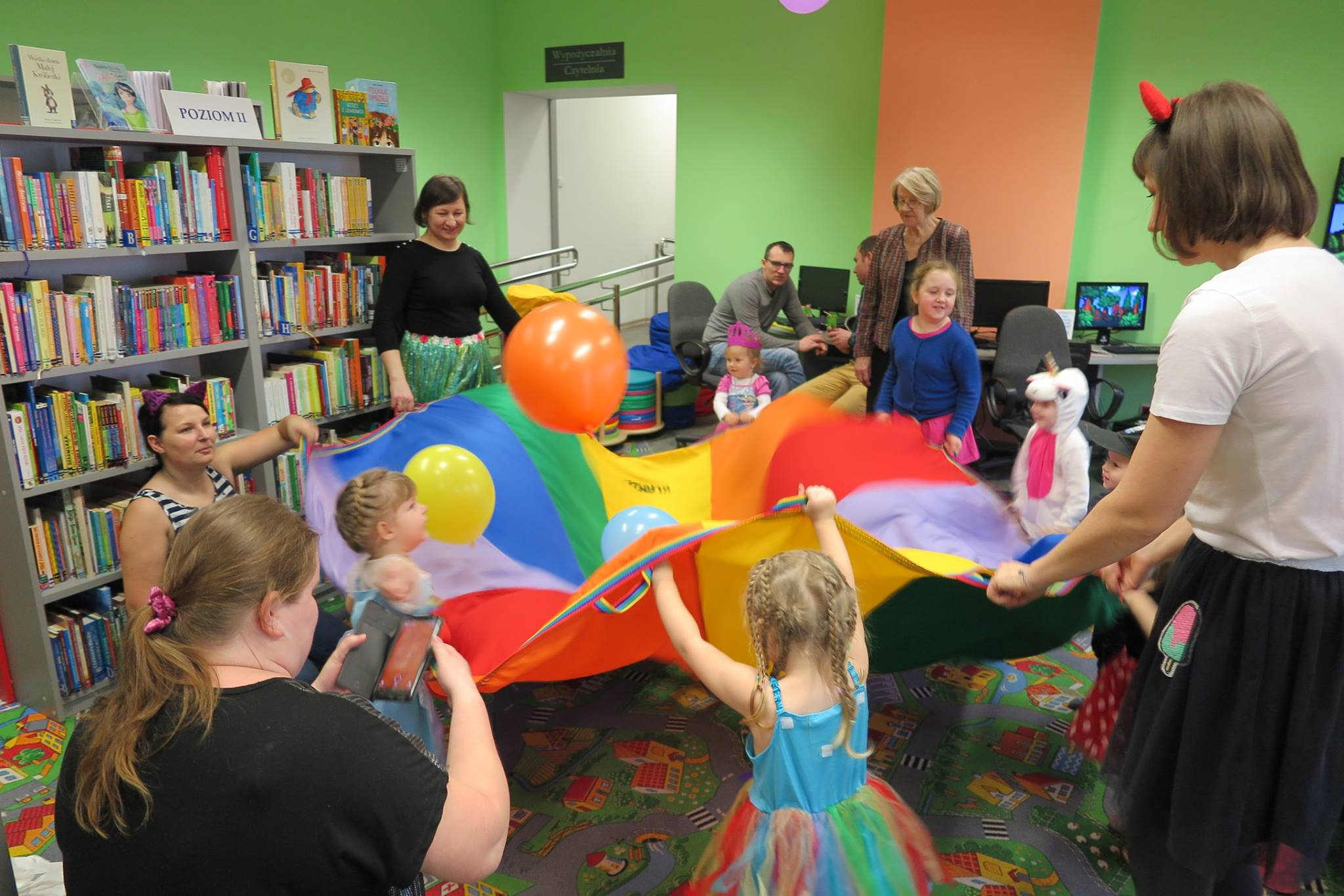 Wnętrze biblioteki. Dzieci wraz  z opiekunami bawią się kolorową chustą na której są kolorowe balony. 