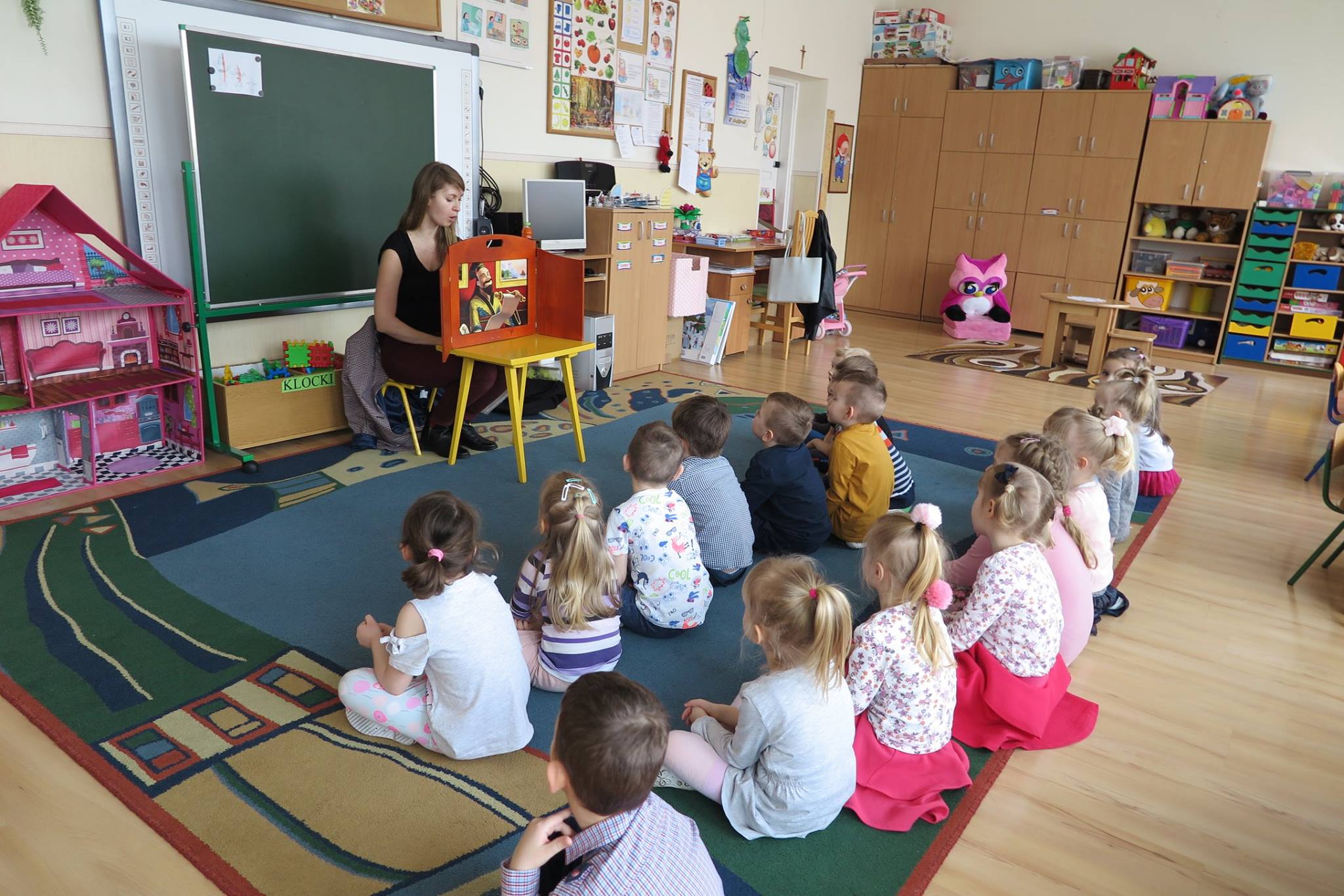 Wnętrze przedszkola nr 1, na kolorowej wykładzinie siedzą dzieci i słuchają teatrzyku  "Słowik",który czyta bibliotekarka Ola Cybulska.