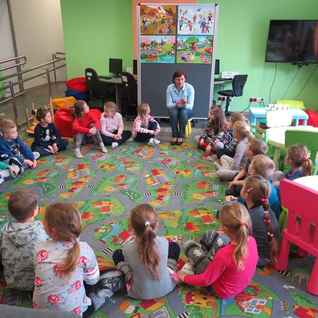 Wnętrze biblioteki. Bibliotekarka Ania Wiśniewska rozmawia z dziećmi o zmieniających się porach roku. Maluchy siedzą w kole na pufach i uważnie słuchają.