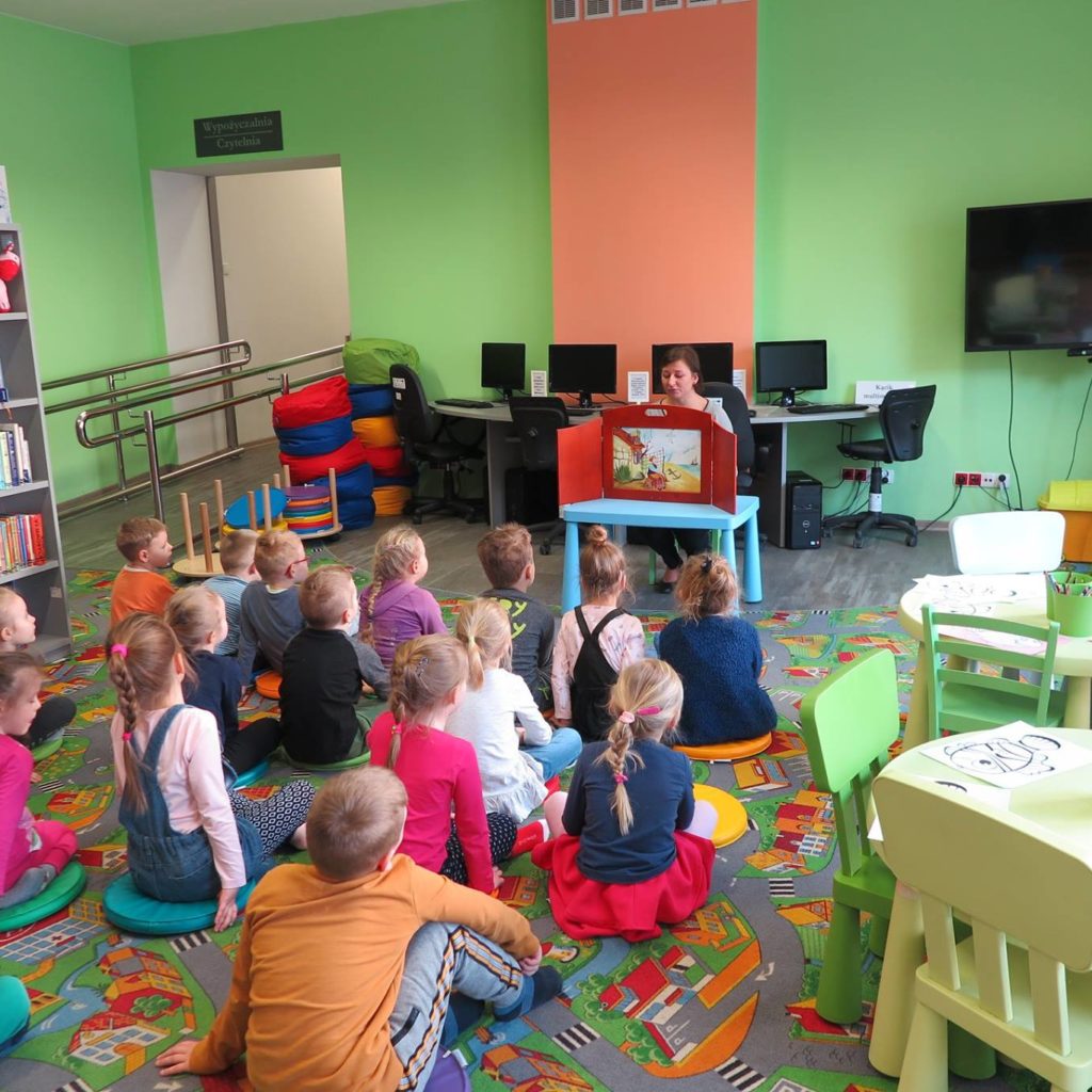 Wnętrze biblioteki. Dzieci słuchają teatrzyku Kamishibai "O rybaku i złotej  rybce", czyta bibliotekarka Ania Wiśniewska.