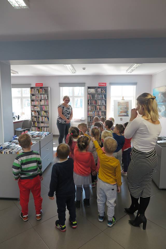 Wnętrze biblioteki. Dzieci zwiedzają mediatekę. Bibliotekarka Dorota Kusik opowiada jakie zbiory można wypożyczyć w mediatece.