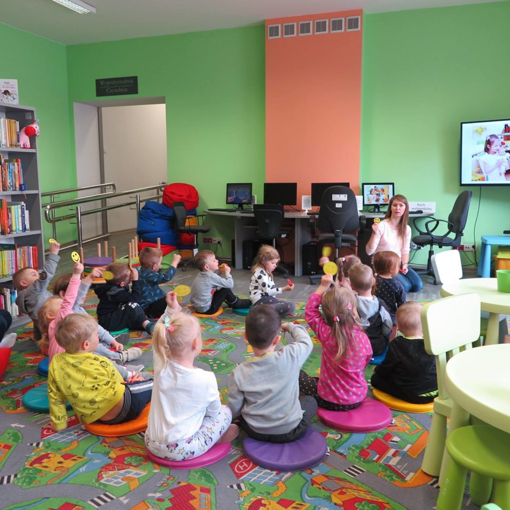 Wnętrze biblioteki. Dzieci siedzą na pufach i biorą udział w lekcji bibliotecznej o ''dzieleniu się''. Zajęcia przeprowadza bibliotekarka Aleksandra Cybulska.