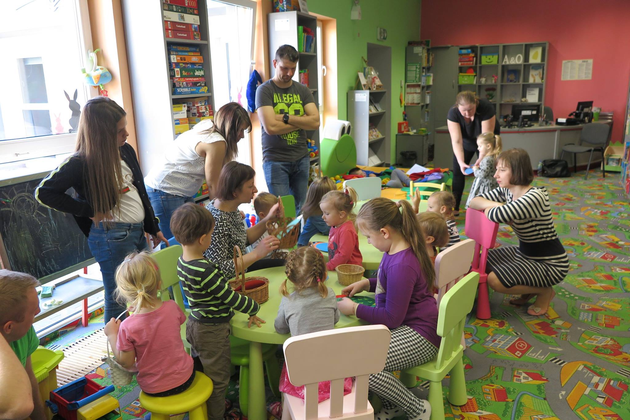 Przy stolikach siedzą dzieci z ''Klubu Malucha''  wraz  z opiekunami i wykonują prace plastyczne. Ozdabiają wycięte jajka kolorowymi paseczkami. Zajęcia prowadzi bibliotekarka Ania Wiśniewska.
