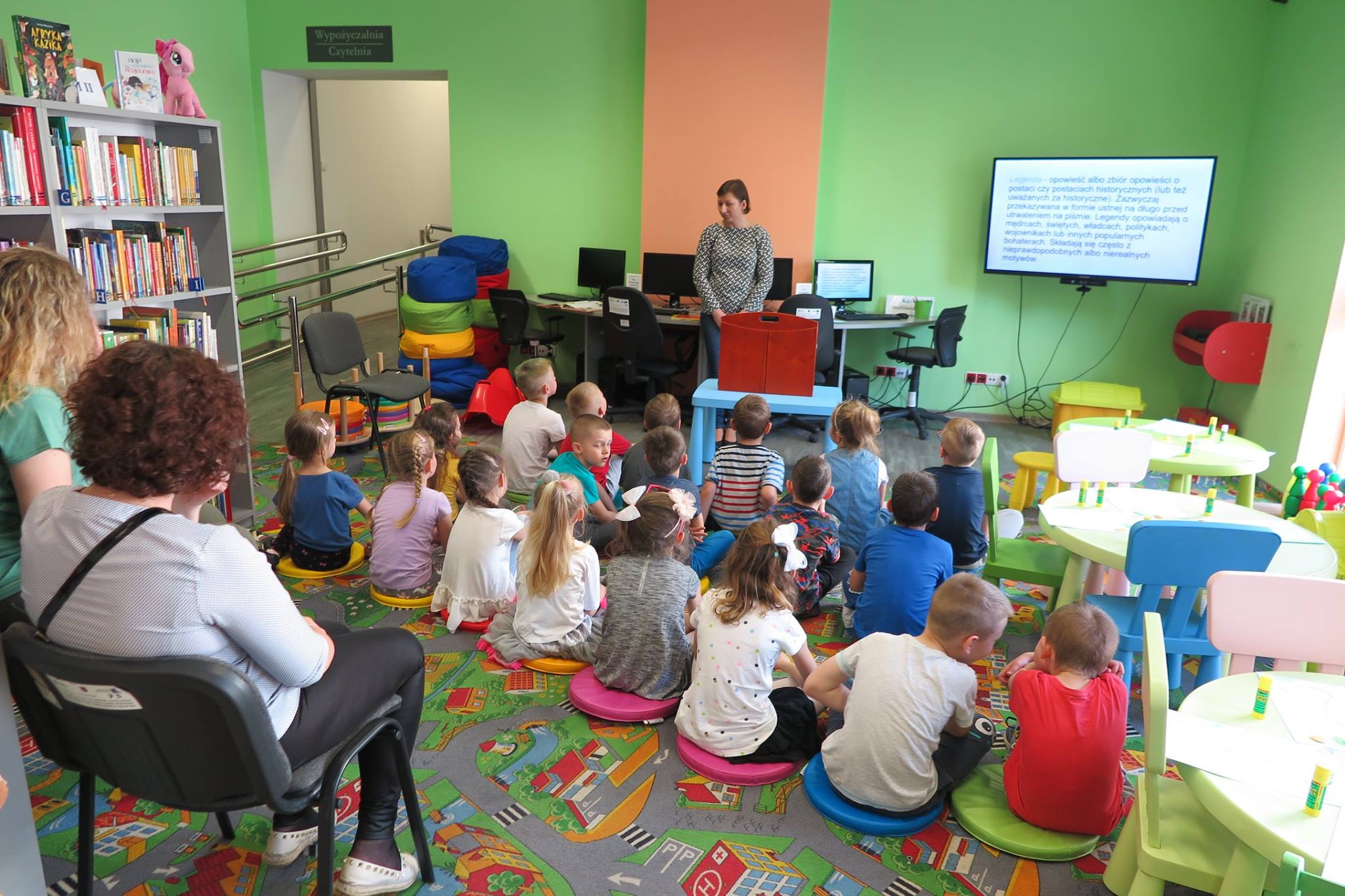 Wnętrze biblioteki. Na pufach siedzą dzieci biorą udział w lekcji edukacyjnej, która miała na celu  przybliżenie polskich legend. Zajęcia prowadzi bibliotekarka Ania Wiśniewska.