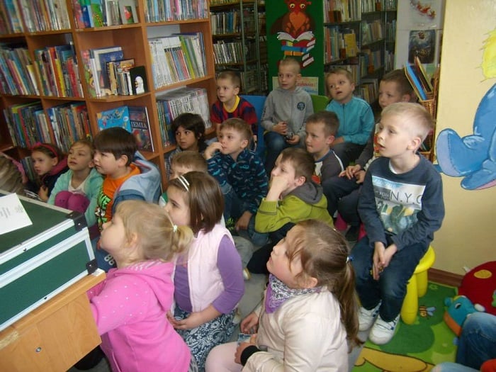 Wnętrze biblioteki, między regałami z książkami siedzi grupa dzieci bierze udział w lekcji bibliotecznej.