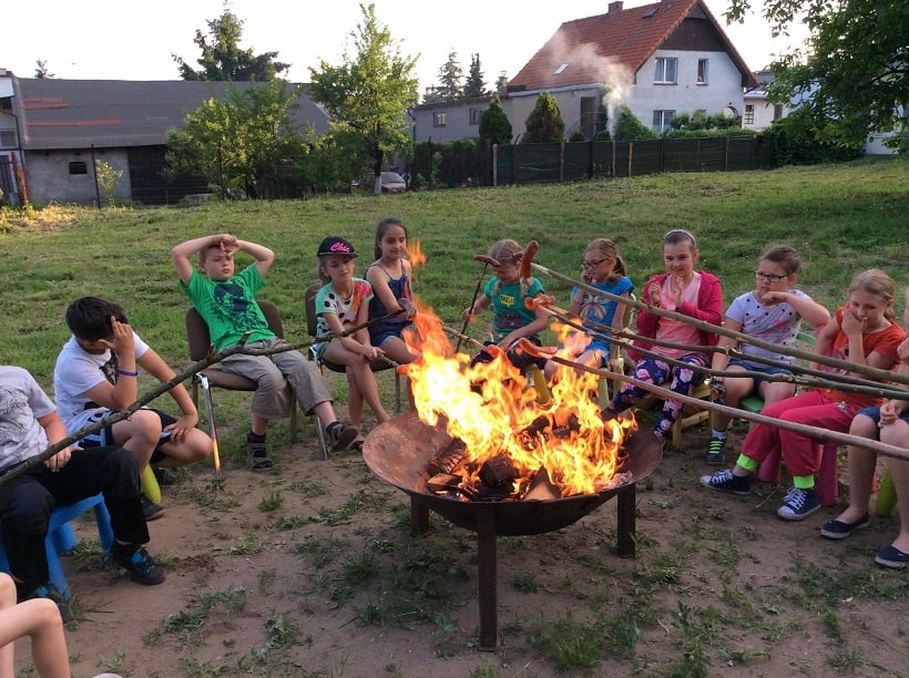 Grupa dzieci siedzi przy ognisku wszyscy trzymają kije na których mają kiełbaski.