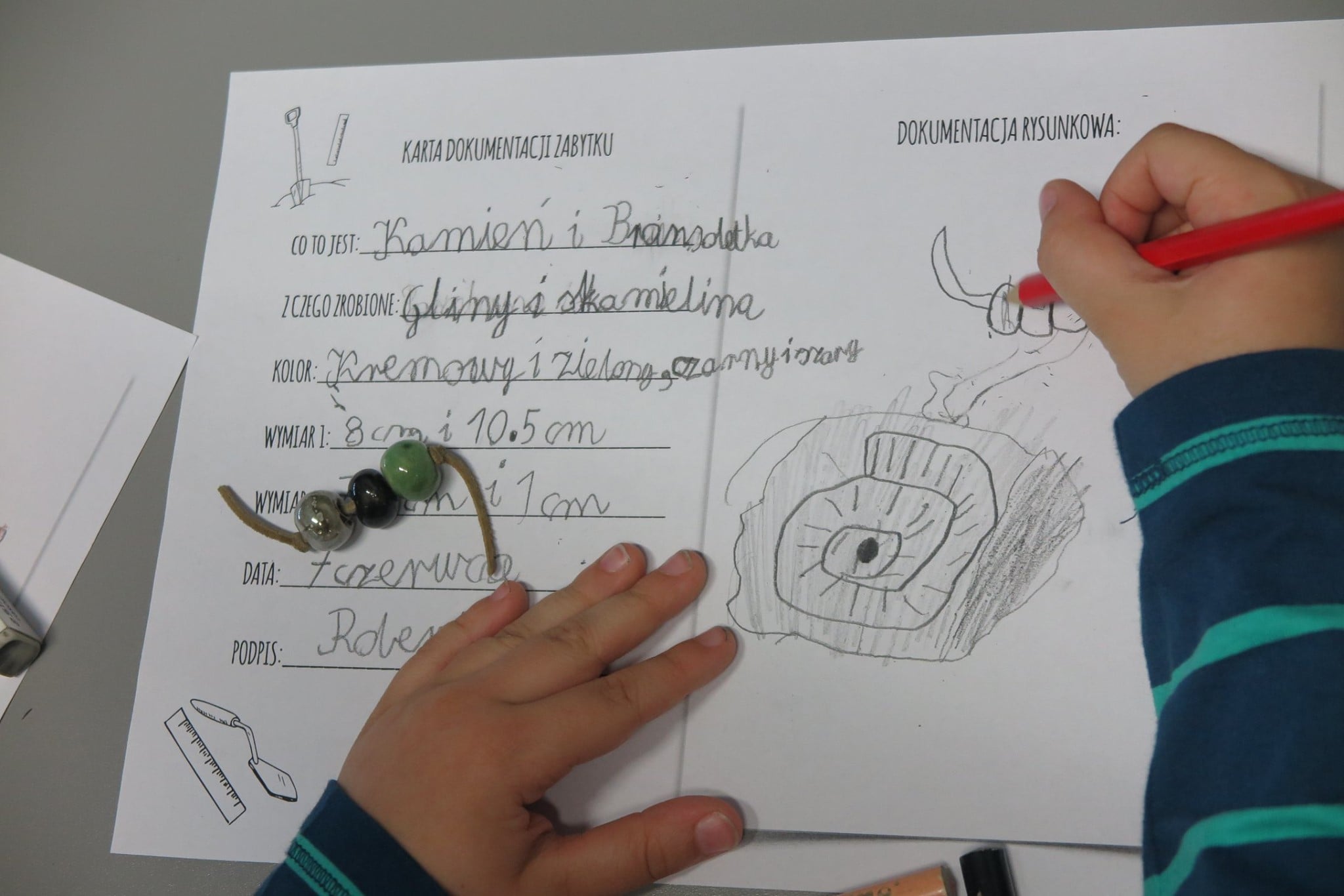 Chłopiec uzupełnia kartę dokumentacji zabytku na drugiej stronie karty wykonuje rysunek kamienia i bransoletki. 