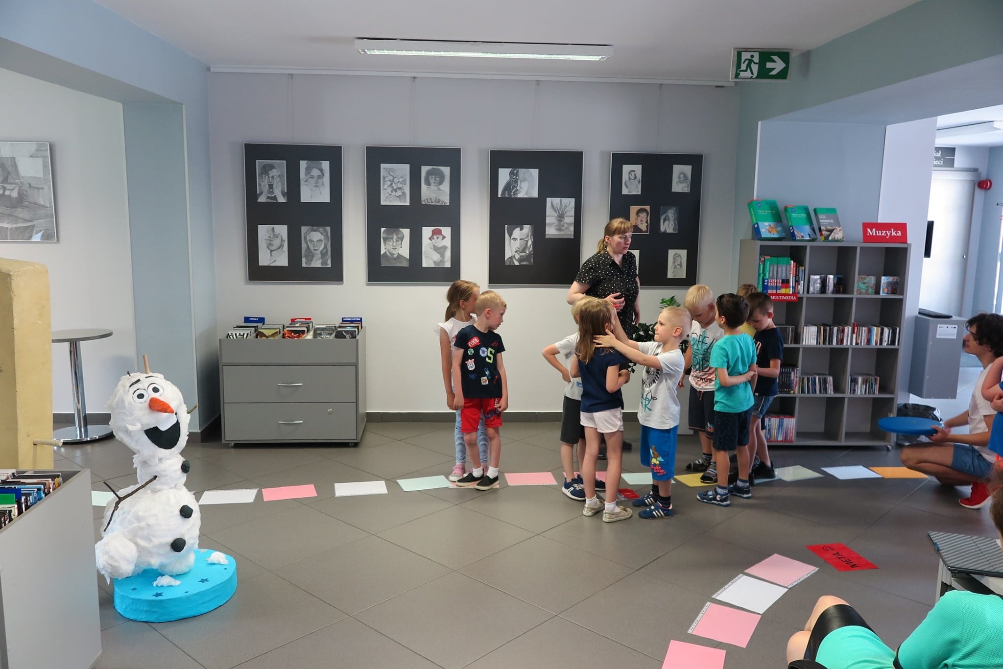 Wnętrze biblioteki, mediateka. Dzieci biorą udział w grze planszowej w ramach obchodów Dni Rodziny. Zajęcia prowadzi bibliotekarka Małgorzata Trąmpczyńska. 