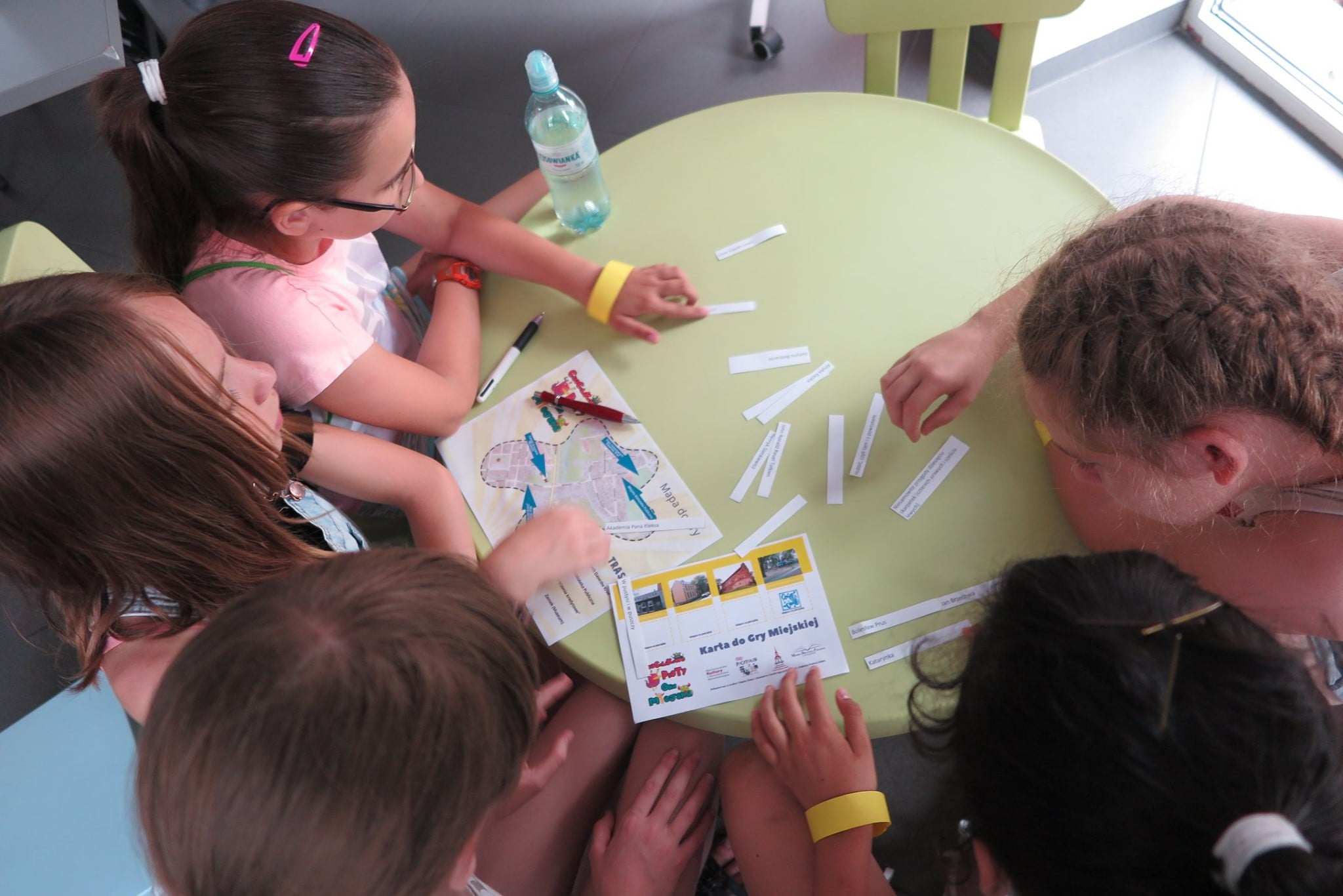 Grupa dzieci przy stoliku rozwiązuje kolejną zagadkę z gry miejskiej „Chochlikowe  psoty”.
