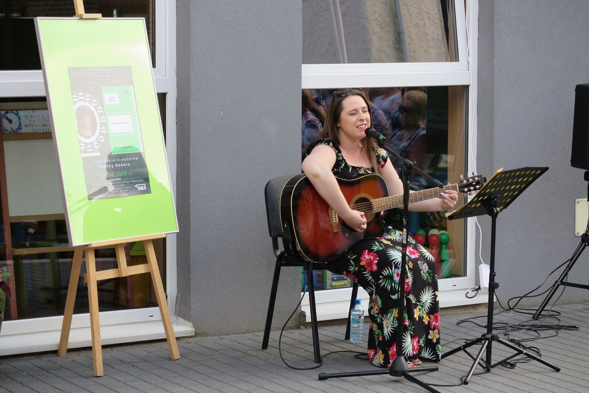 Na placu śpiewa i gra na gitarze Marta Wiadrowska obok sztaluga z plakatem ''Spotkanie z autorką Janiną Dzenis''. 