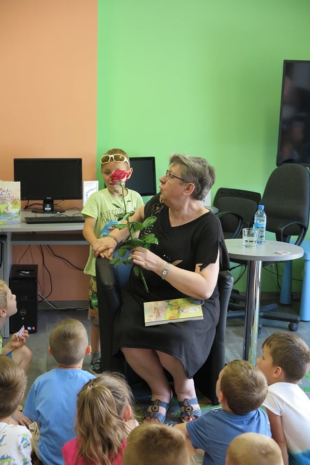 Wnętrze biblioteki. Na fotelu autorka Ewa Sotomska. Chłopiec w imieniu wszystkich dzieci wręcza różę w podziękowaniu za spotkanie. 