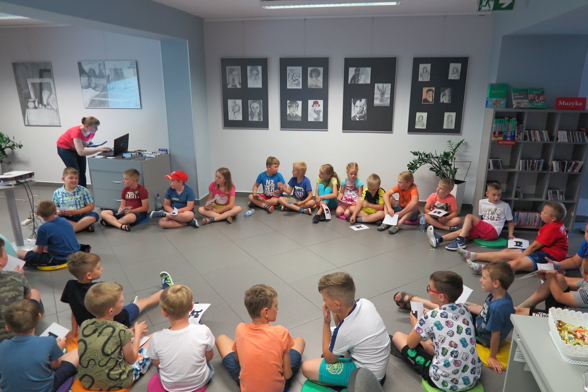 Wnętrze biblioteki, mediateka.  Na pufach siedzą dzieci biorą udział w zajęciach z programowania z Minecrafta Education Edition. Zajęcia prowadzi bibliotekarka Małgorzata Trąmpczyńska.