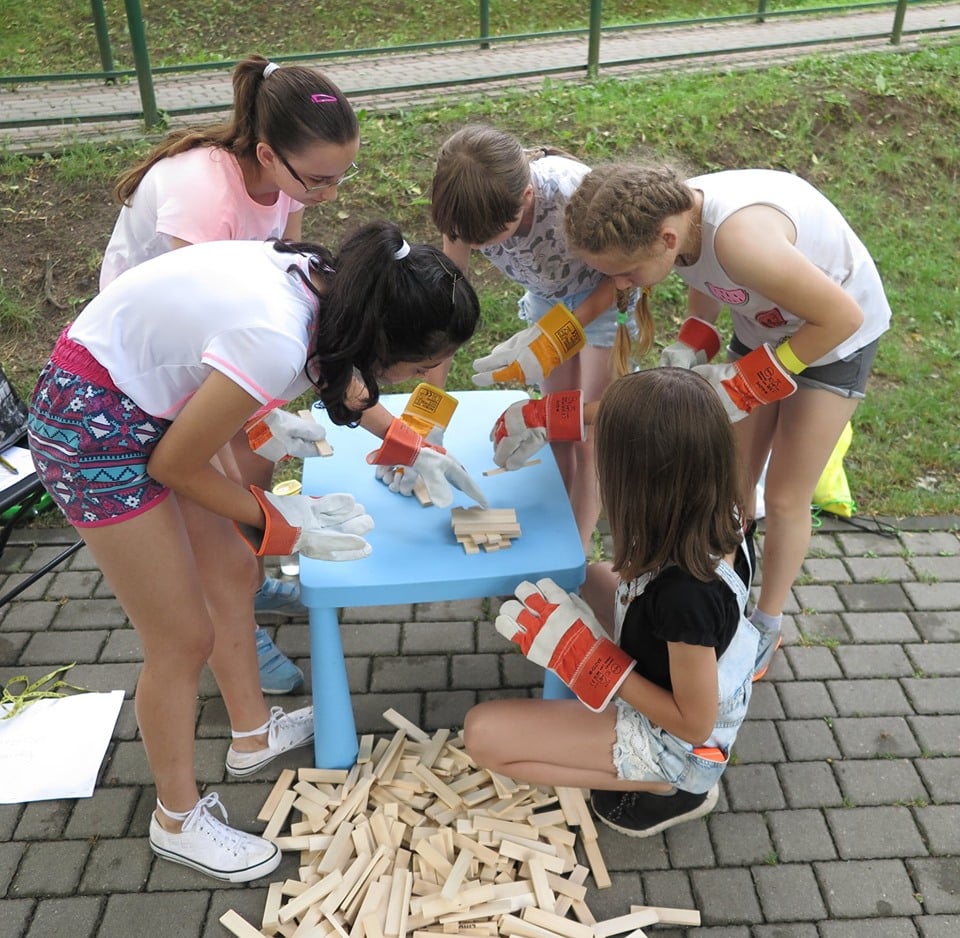 Grupa dzieci przy stoliku układa drewniane klocki wykonuje zadanie z gry miejskiej „Chochlikowe  psoty”.