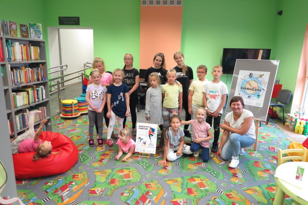 Wnętrze biblioteki. Grupa dzieci z bibliotekarką Anią Wiśniewską biorąca udział w warsztatach tanecznych. 