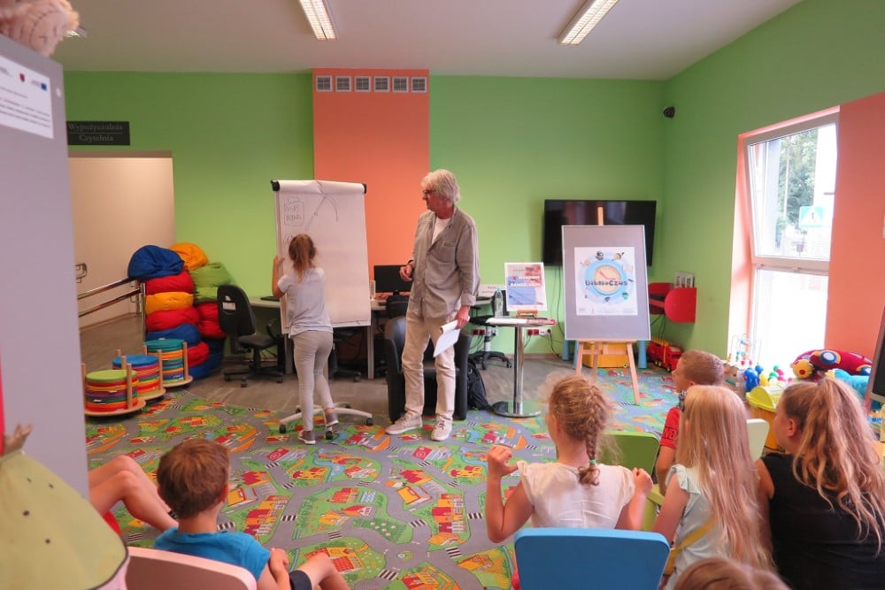 Wnętrze biblioteki. Oddział dla dzieci. Dziewczynka bierze udział w zajęciach dla dzieci które prowadzi Marek Samselski.