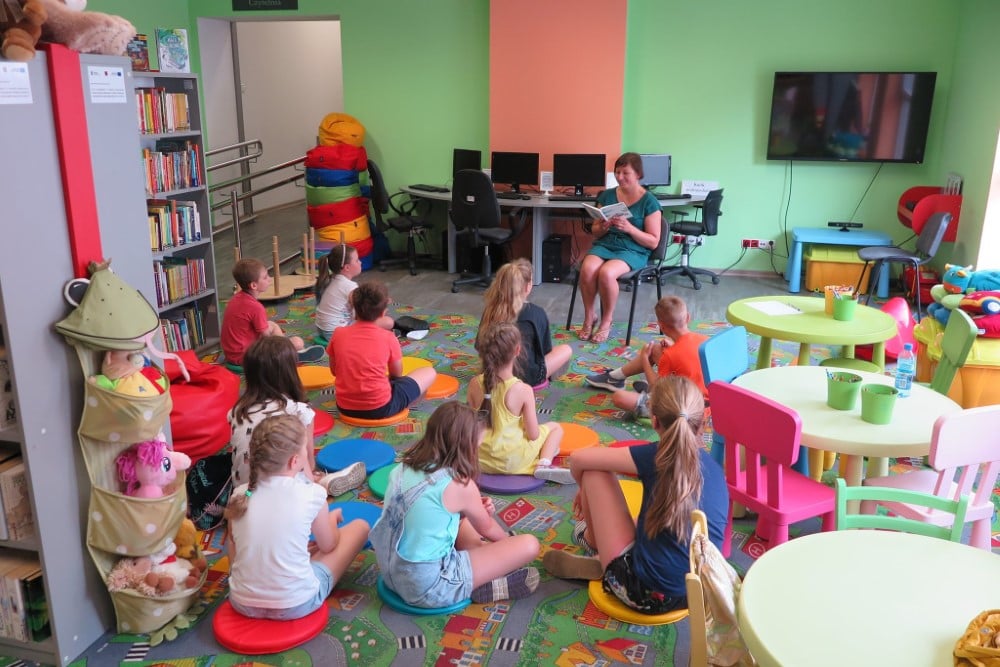 Wnętrze biblioteki. W pierwszym tygodniu wakacji dzieci słuchają opowiadania. Czyta bibliotekarka Ania Wiśniewska.