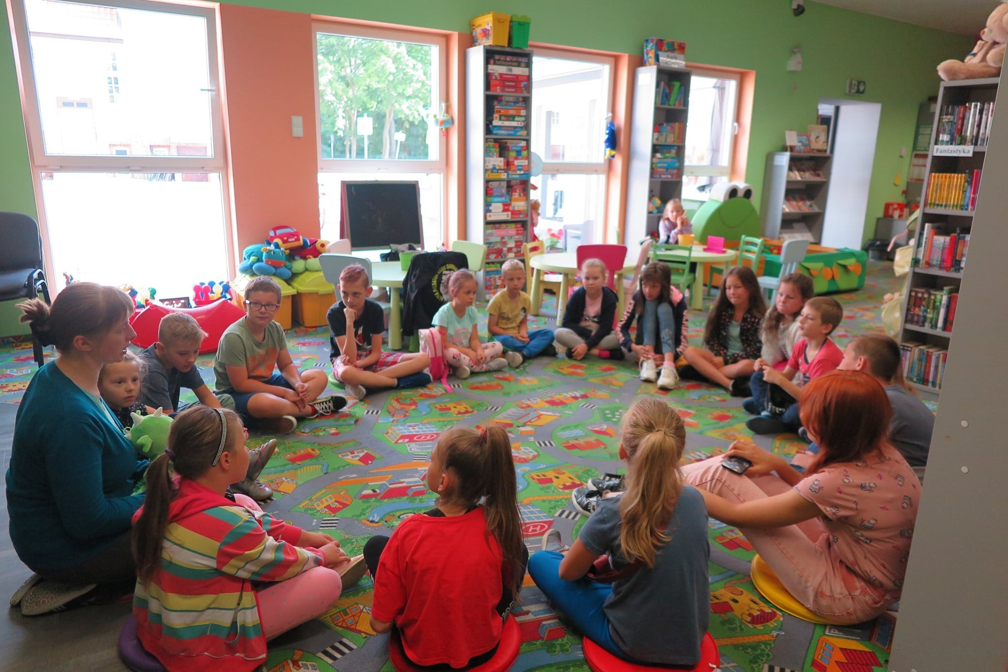 Wnętrze biblioteki. Dzieci siedzą w kole i słuchają opowiadania. Zajęcia prowadzi bibliotekarka Małgorzata Trąmpczyńska.
