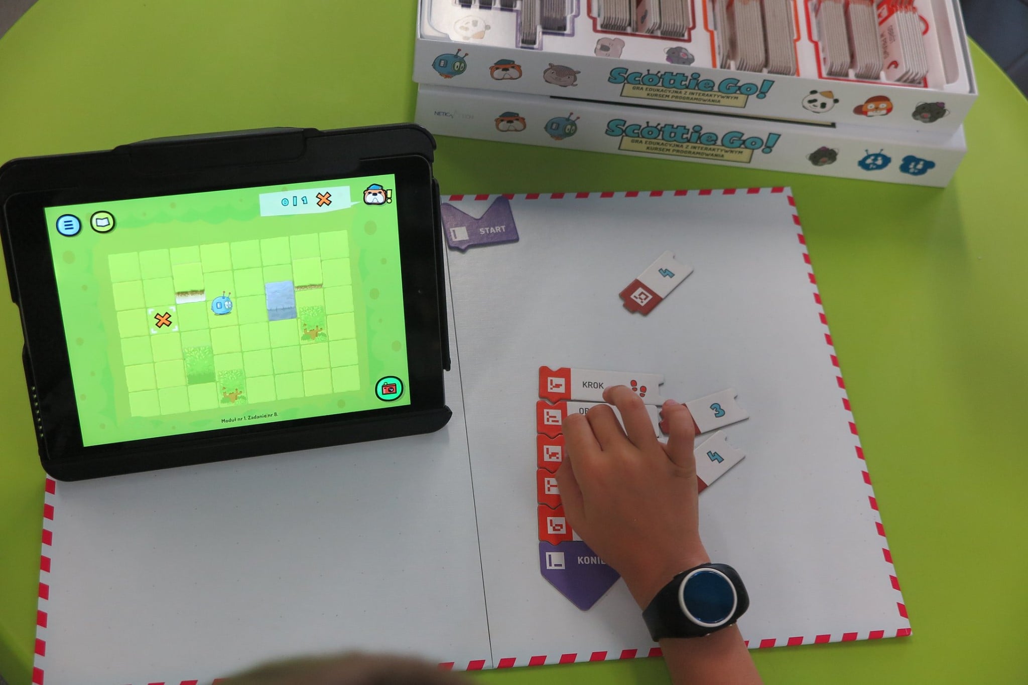 Na stoliku gra z programowania ''Scottie Go'' i tablet na którym gra chłopiec.