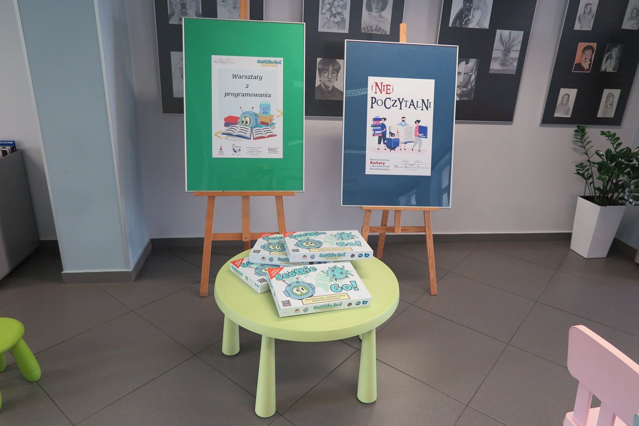 Dwie sztalugi z plakatami, od lewej plakat ''Warsztaty z programowania'' i plakat ''(Nie) poczytalni'' na zielonym stoliku leżą 4 gry ''Scottie Go''. 