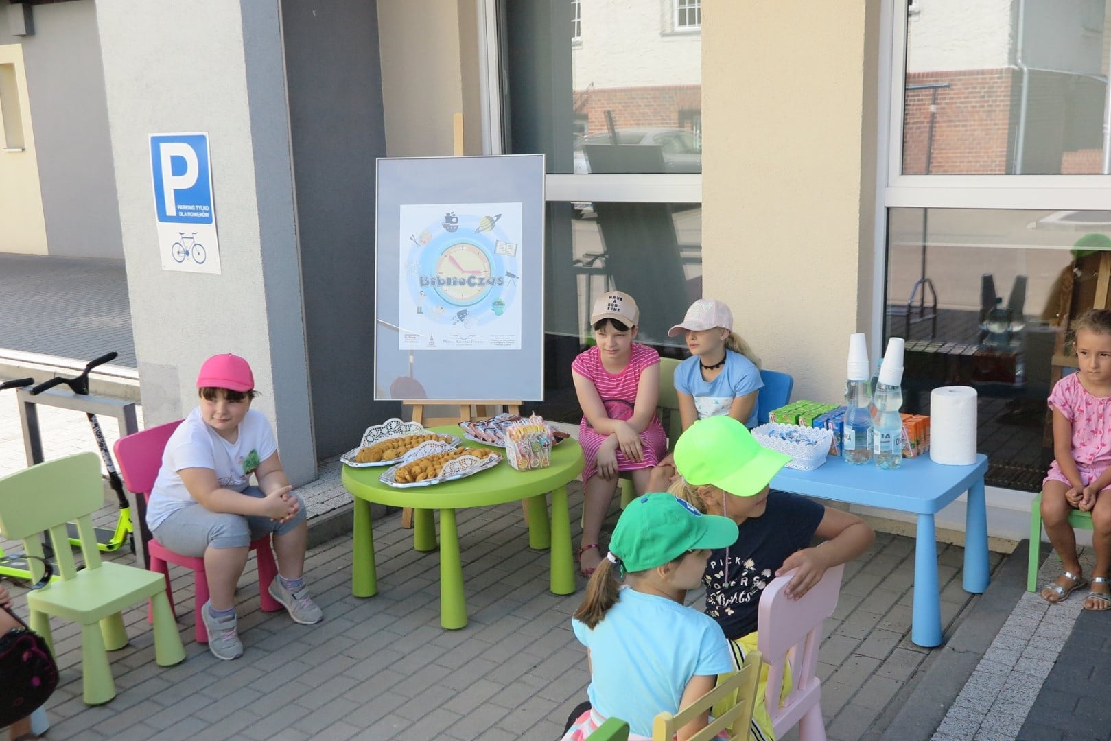 Na placu MBP przy kolorowych stolikach siedzą dzieci, na stolikach ciastka, soczki i woda dla dzieci. 