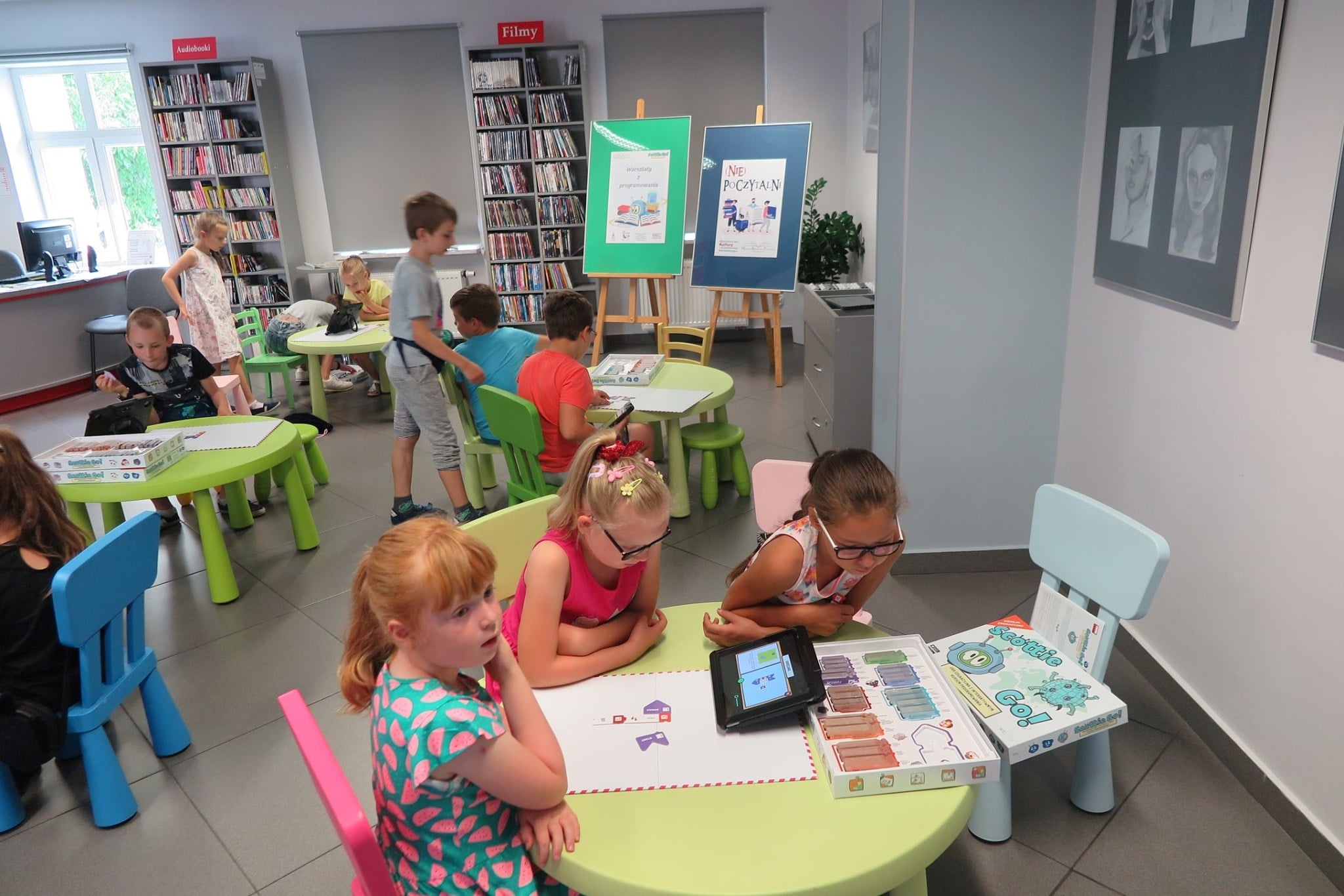 Wnętrze biblioteki,mediateka. Przy stolikach dzieci grają w grę z programowania ''Scottie Go''.