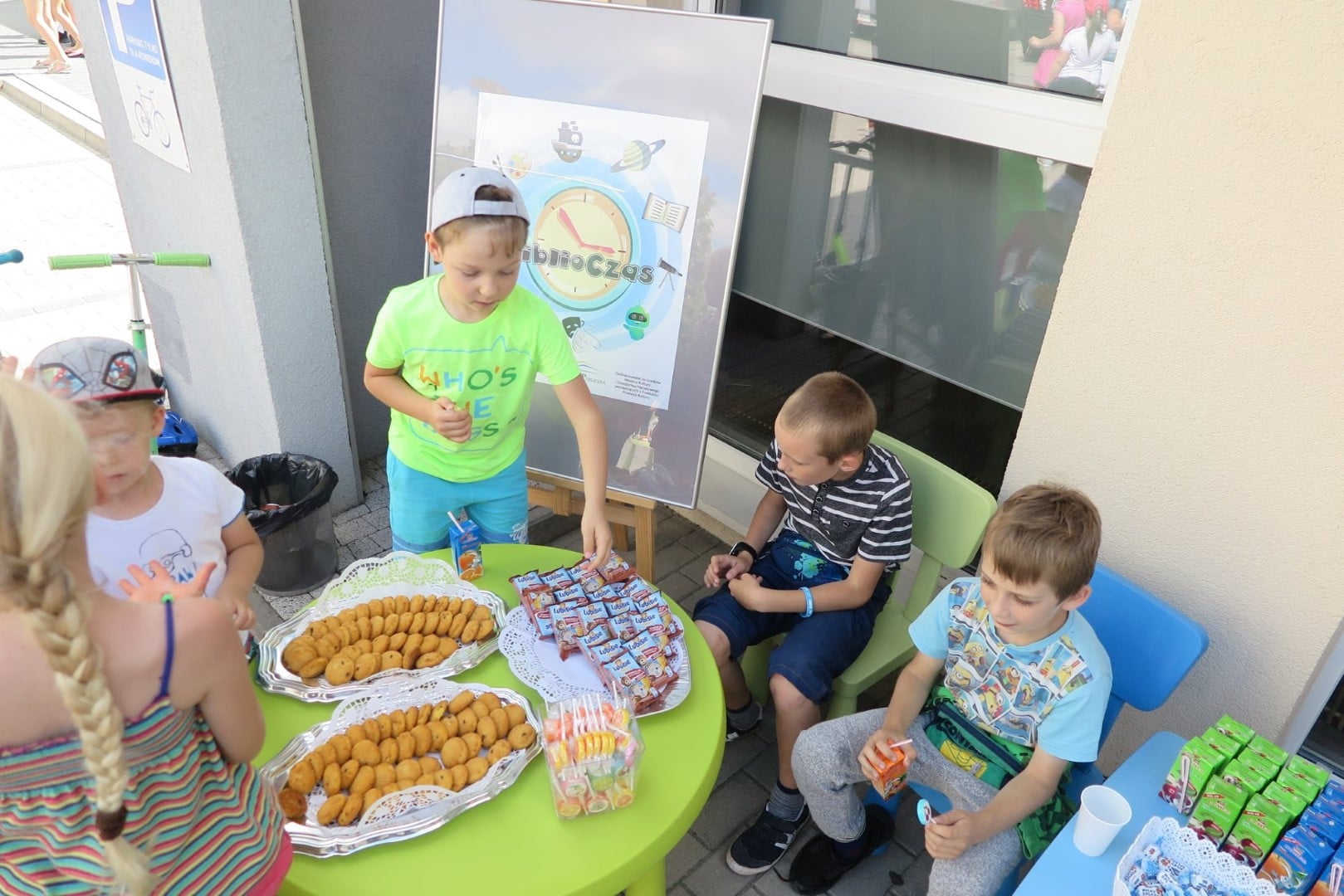 Na placu MBP przy zielonym stoliku dzieci jedzą ciastka i piją soki. 