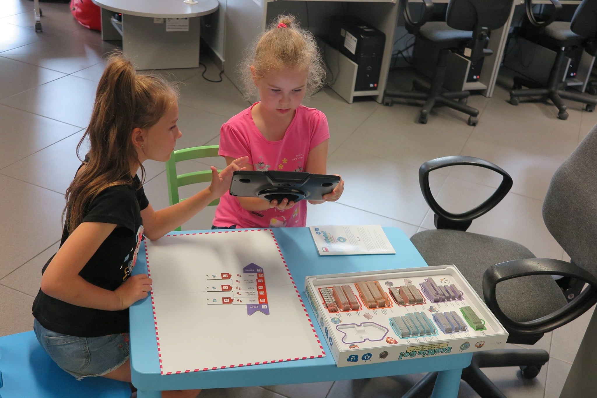 Wnętrze biblioteki,mediateka. Przy stoliku dwie dziewczynki grają w grę z programowania ''Scottie Go''.