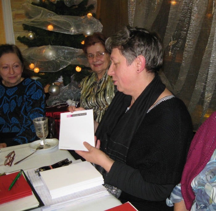 Przy stolikach siedzą członkowie DKK UTW wspólnie omawiają książkę E. Sukertowej-Biedrawiny "Diabeł na Mazurach w baśniach i podaniach", w tle choinka świąteczna.