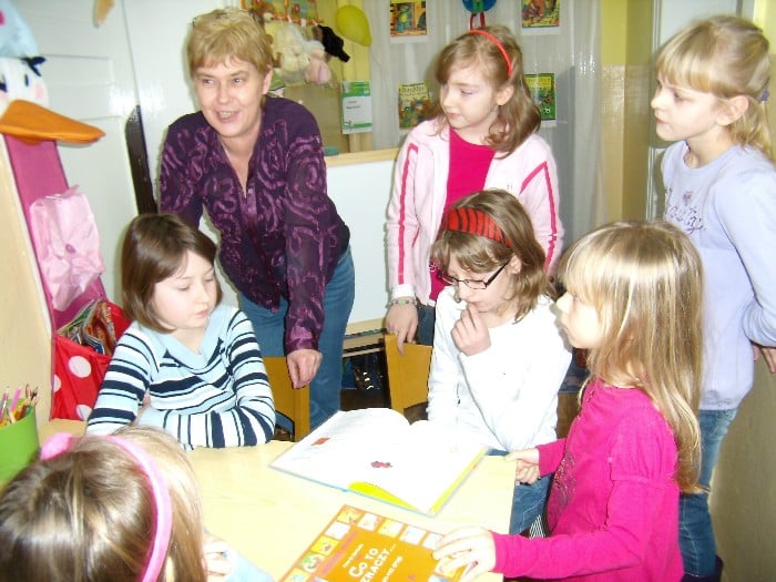 Wnętrze biblioteki. Przy stoliku sześć dziewczynek czytają razem książkę obok bibliotekarka Marzena Pawłowska.