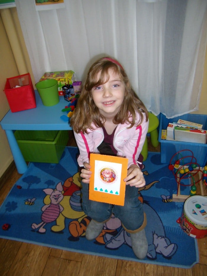 Dziewczynka trzyma wykonaną przez siebie kartkę z życzeniami dla rodziców.